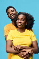 Obraz premium Cheerful young African couple embracing each other against a bright blue background, dressed in colorful yellow and orange shirts, showcasing love and connection