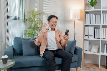A young man sits comfortably on a sofa with headphones around his neck, smiling and waving while enjoying a video call on his smartphone, creating a relaxed and friendly scene.