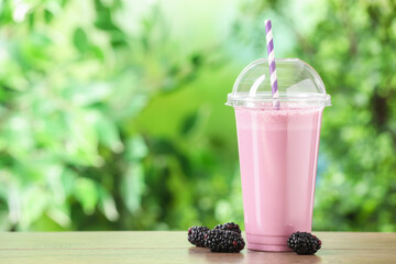 Tasty milkshake in plastic cup and blackberries on wooden table outdoors, space for text
