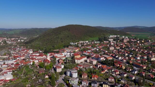 Scenic aerial flight above the residential town of Tisnov, South Moravia, Czechia, showcasing green Boskovice Furrow hills and rooftops on a bright spring day.