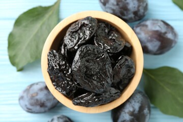 Dried prunes in bowl, leaves and fresh plums on light blue wooden table, flat lay