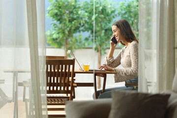 Happy woman talking on phone checking laptop in a terrace