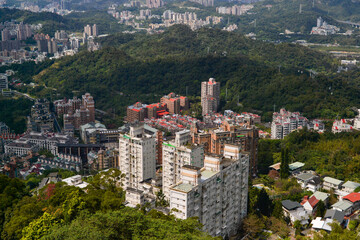 Aerial view of a densely populated city with numerous residential buildings nestled into lush green mountains and hills under a bright sky, showcasing vibrant urban development. Cityscape.