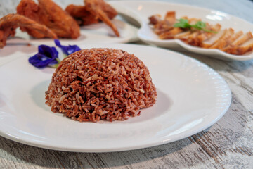 Mound of healthy brown rice on a white plate with crispy fried chicken wings and sliced pork belly in the background, promoting Asian cuisine and healthy eating.
