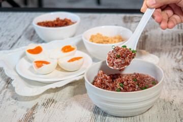 Adult hand serving brown rice porridge with chives into a white bowl, accompanied by salted duck eggs and minced pork on a rustic wooden table, Asian Cuisine.