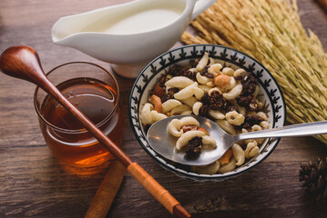Bowl of mixed nuts, granola, and dried fruit with honey, milk, and wheat stalks on a rustic wooden table, healthy breakfast concept.