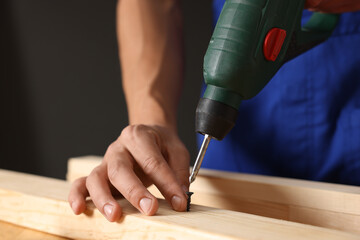 Professional repairman twisting screw into wooden plank with electric screwdriver indoors, closeup