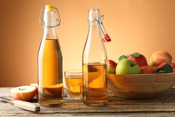 Delicious cider in bottles, glass, apples and knife on wooden table against orange gradient background