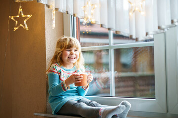 Little preschool girl holding cup with hot chocolate with marhsmallows. Happy child drinking sweet cocoa by window with Christmas lights in winter. Cozy family celebration of xmas.