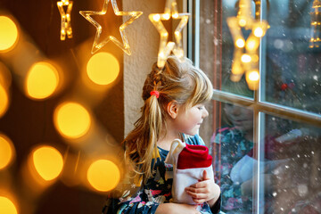 Little preschool girl holding cup Santa Claus boot with gift called Nikolausstiefel in German. Happy child wait on holiday by window with Christmas lights in winter. Cozy family celebration of xmas.