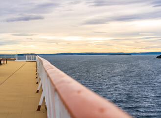 Golden hues fill the sky as the sun sets over Oslofjord in Norway. A ship's railing lines the deck, offering a scenic view of the calm waters and distant islands during a warm summer evening sunset