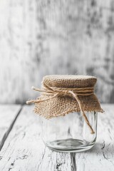 Glass jar with burlap cover and rope bow on white wooden table with rustic background in natural light