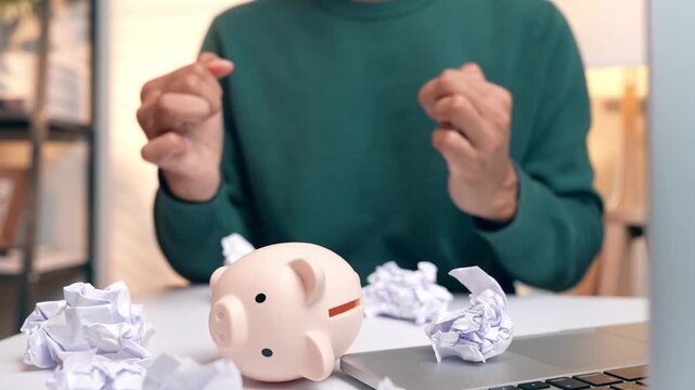A stressed person crumpling paper with a fallen piggy bank, symbolizing debt burden and financial failure, while also reflecting frustration, lack of savings stability, and emotional strain.
