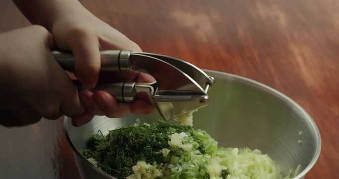 Close-up of a cook's hands pressing garlic cloves into a special garlic press to prepare a filling for baked goods. Chopping garlic into a container with dill and green onions.