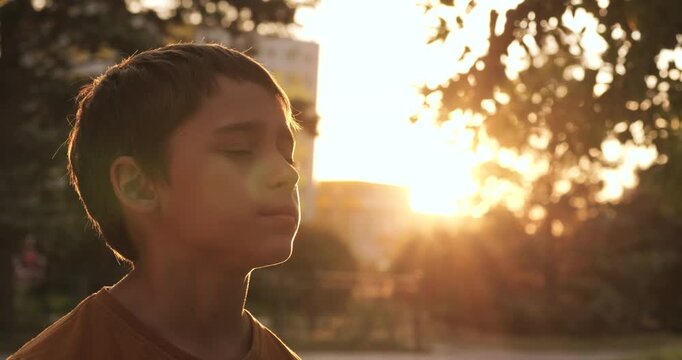 Boy in sunset park stands with eyes closed. Face turns to warm light, slow breathing shows calm. Soft flare glows around profile. Nature background creates peaceful mood. wellness content