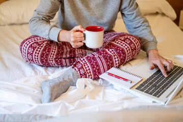 Woman sitting comfortably in bed, wearing cozy pajamas and warm socks, holding a steaming mug while typing on her laptop, with a notepad and pen at her side, enjoying a relaxing morning