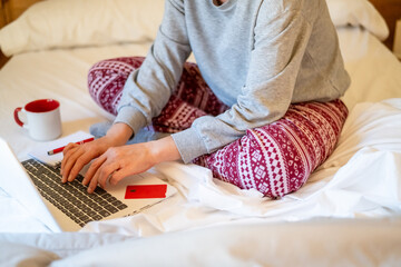 Woman wearing festive christmas pajamas sits comfortably on a cozy bed, using a laptop while enjoying a warm cup of beverage, with a credit card and notepad nearby for online shopping