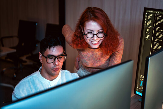 Two coworkers collaborate at dual monitors in a dim office, reviewing code and brainstorming solutions during late night software development.
