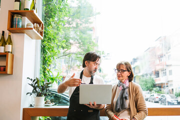 Cafe barista and customer reviewing information on a laptop by a large window, discussing business ideas in a bright urban coffee shop with plants and street view.