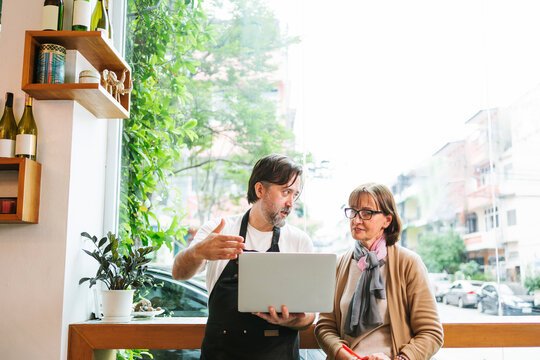 Cafe barista and customer reviewing information on a laptop by a large window, discussing business ideas in a bright urban coffee shop with plants and street view.