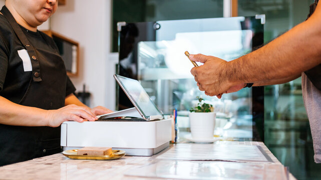 Customer uses smartphone for contactless payment at a retail counter as a cashier operates the POS terminal behind a protective screen in a cafe setting.