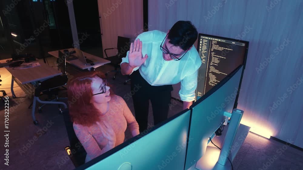 Poster two coworkers celebrate a coding win with a high five in a modern office, working at dual monitors w