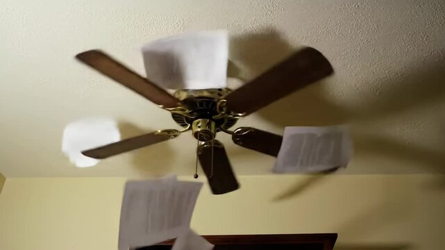 Indoor shot of a ceiling fan spinning, blowing white papers around the room, brown blades, home interior, appliance, cooling system, air circulation.