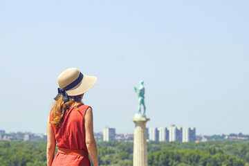 Back view of stylish woman in dress a hat exploring Belgrade fortress. Female tourist near Pobednik monument. Concept of slow travel, inspiration and mindful tourism in historic European cities.