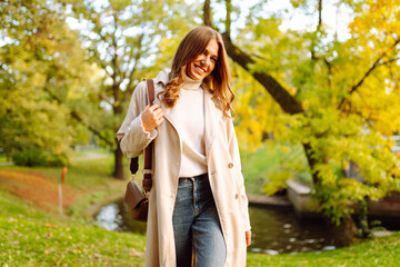 A joyful woman in a stylish trench coat poses in an autumn park. A beautiful woman enjoys a walk outdoors. Beauty and fashion concept. Autumn mood.