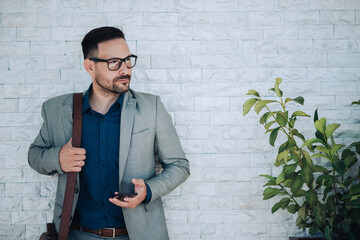 Obraz premium Businessman holding smartphone and looking away next to white wall