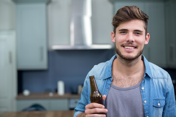 Adult man holding beer bottle and standing in home kitchen with grey cabinets, utensils, copy space