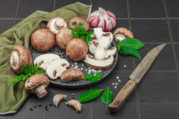 Fresh brown mushrooms with garlic, herbs and rustic knife on black tile kitchen surface