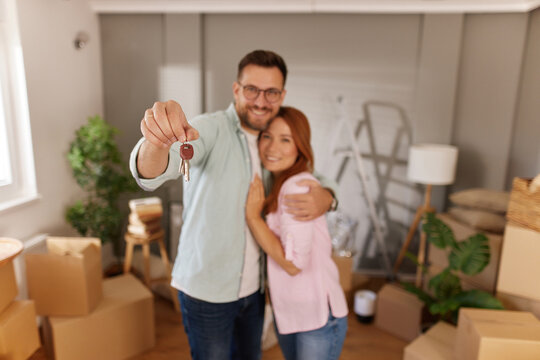 A couple joyfully embraces while holding keys to their new home, surrounded by packed boxes in a bright and inviting living space filled with greenery.