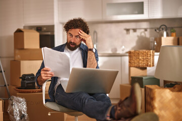 A man sits in a chair surrounded by moving boxes, looking stressed while reviewing documents on his laptop. He appears to be managing the chaos of relocating.