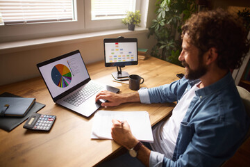Fototapeta premium A man is engaged in work at his home office, using a laptop and tablet to analyze data. His workspace is organized with notes, a calculator, and houseplants, enhancing productivity.