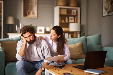 A man expresses concern over financial issues while a woman comforts him in a cozy living room. They sit together on a couch with papers and a laptop nearby, reflecting their struggles.