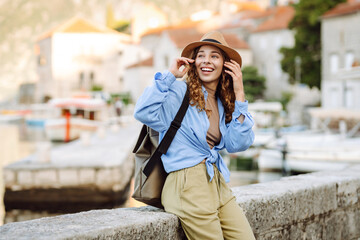 Fototapeta premium Portrait of a young woman wearing a hat and carrying a briefcase strolling along the sea in a picturesque town. The traveler enjoys the seascape. Concept of tourism and enjoyment.