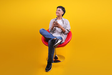 Joyfully Young Asian Male Using Smartphone While Relaxing Sitting On Modern Chair