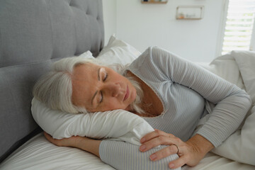 Mature woman lying on side in bedroom showing white pillow comforter grey headboard blinds