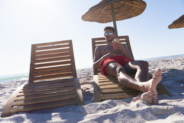 Man reclining on wooden lounge chair under straw parasol at beach with cocktail glass, copy space