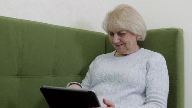 Smiling senior woman in gray knitted sweater using black tablet on green sofa, symbolizing digital learning and online communication.