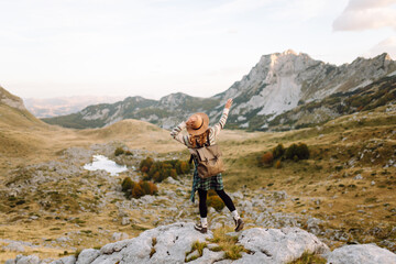 Fototapeta premium A young female hiker with a backpack and hat sits on a rock, looking at the mountain scenery. A woman on a hiking trail feels free and enjoys the adventure. Concept of travel, freedom.