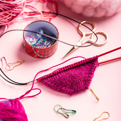 Colorful knitting supplies are beautifully displayed on a soft pink surface, including yarn, needles, scissors, and stitch markers, emphasizing the joy of crafting
