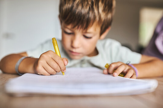 Young boy writing with yellow pen in notebook while studying and doing homework indoors
