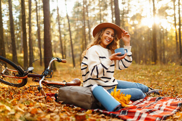 A happy woman sits on a red blanket next to a bicycle, drinking hot tea from a thermos. A young woman relaxes in a clearing in an autumn park. Picnic and cycling concepts