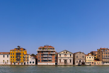 Colorful Venetian waterfront buildings under clear sky. Row of vibrant historic buildings along the waterfront in Venice, Italy, viewed from across the canal on a sunny, cloudless day.