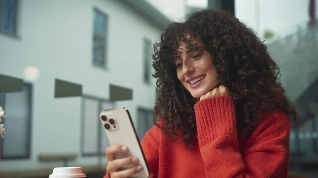 Caucasian woman with curly hair smiling while looking at smartphone in hand, sitting indoors near window. Female in red sweater drinking takeaway coffee and browsing phone at cafe table. - Powered by Adobe