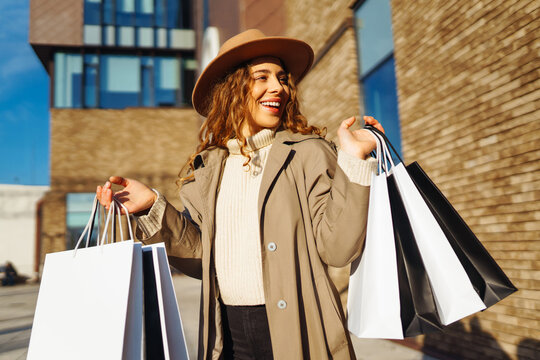 Portrait of a smiling woman in a hat holding shopping bags on a sunny day. A young woman strolls through a busy city after shopping. Concept of discounts, weekend.
