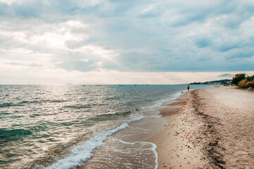 Scenic view of a lone man walking along the sandy beach of Polychrono in Halkidiki, Greece. Gentle waves meet the shore under a dramatic cloudy sky, fishing boats visible on the horizon.