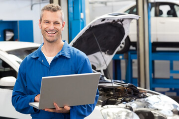 Male mechanic wearing blue coveralls using laptop inspecting lifted car with raised hood in garage